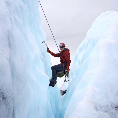 person in red jacket and helmet holding ice tools in glacier crevasse